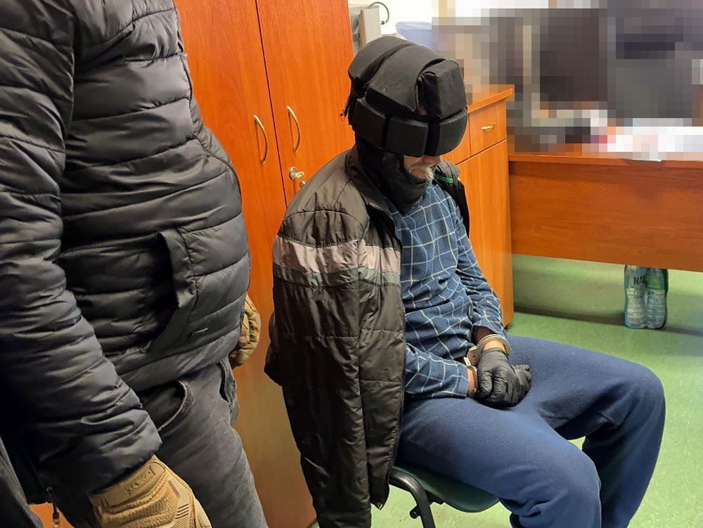Mężczyzna w rękawiczkach siedzący na krześle z zawiązanymi oczami, otoczony przez dwóch ubranych na czarno.