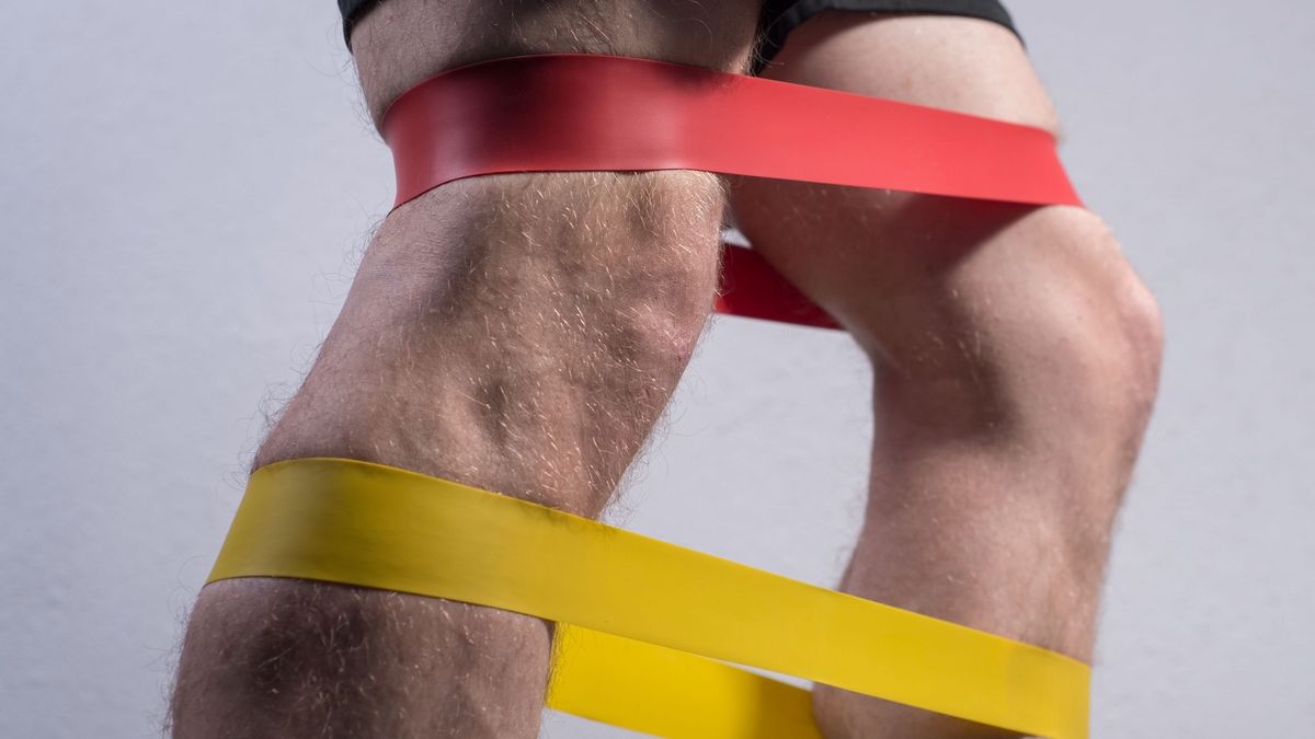 closeup, bottom view of the guys muscular legs in sports shorts, stretching fitness elastic bands with his legs, involvement of leg muscle groups that are not involved in everyday life.closeup, bottom view of the guys muscular legs in sports shorts, stretching fitness elastic bands with his legs, involvement of leg muscle groups that are not involved in everyday life. On a gray background. Advertising fitness gum. Athletic guy. Pronounced muscles of the legs.alexstockphoto21
