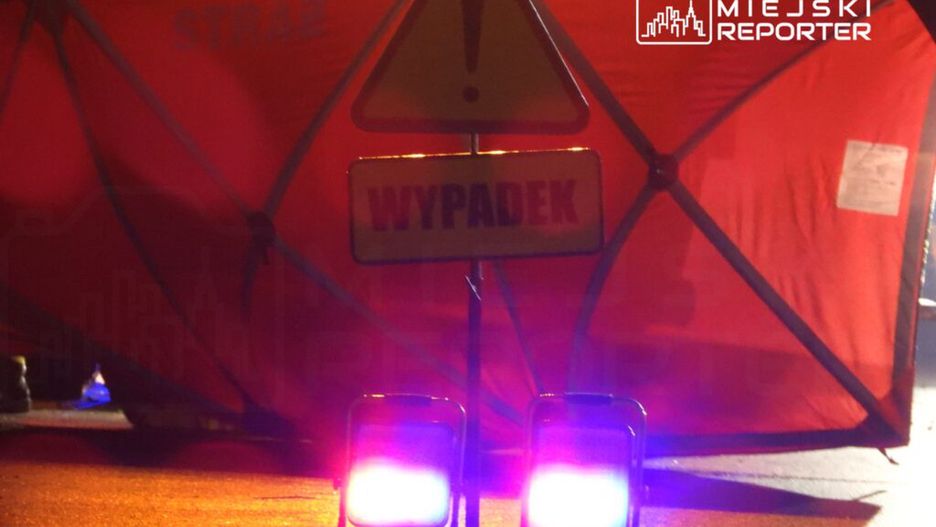 Na drodze stoi znak ostrzegawczy "WYPADek" z migającymi światłami, w tle widoczny czerwony namiot.