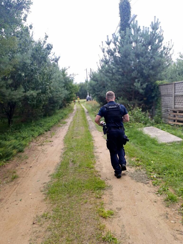 Policjant w mundurze idzie wzdłuż leśnej drogi, mijając drzewa i ogrodzenie.