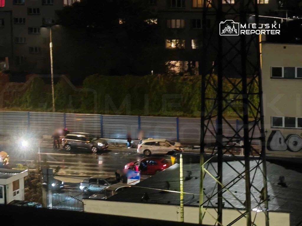 Grupa osób wokół dwóch uszkodzonych samochodów na mokrej nawierzchni, w tle oświetlone budynki.