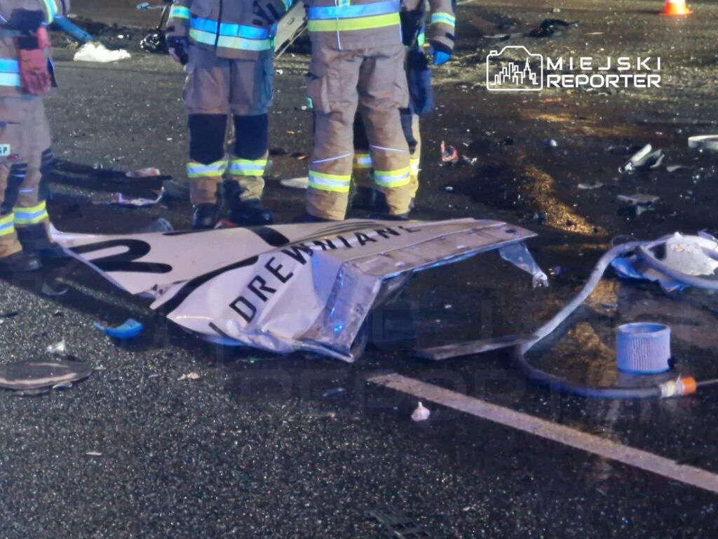Ratownicy w mundurach stoją nad zniszczonym znakiem drogowym na zatarasowanej jezdni po wypadku.