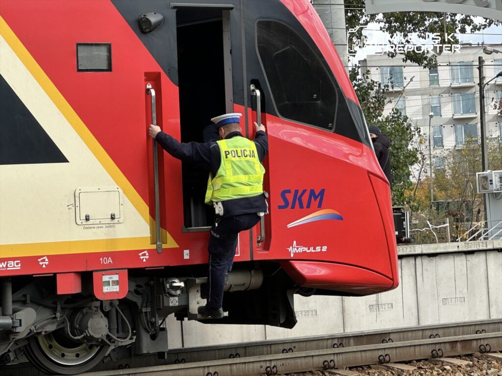 Policjant w kamizelce odblaskowej wchodzi do kabiny czerwonego pociągu SKM na stacji kolejowej.