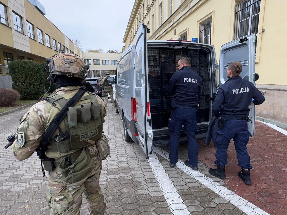 Policjant w mundurze taktycznym obserwuje dwóch funkcjonariuszy policji przy otwartej furgonetce z kratami.