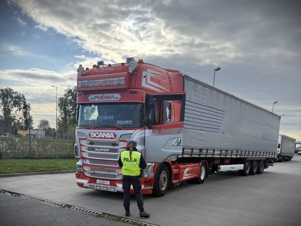 Policjant w kamizelce odblaskowej kontroluje ciężarówkę marki Scania na parkingu w pobliżu drogi.