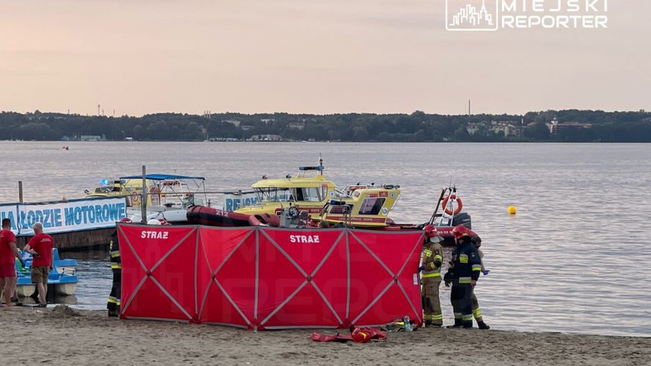 Grupa ratowników w czerwonych koszulach przygotowująca sprzęt na plaży obok wody, z łodziami ratunkowymi w tle.