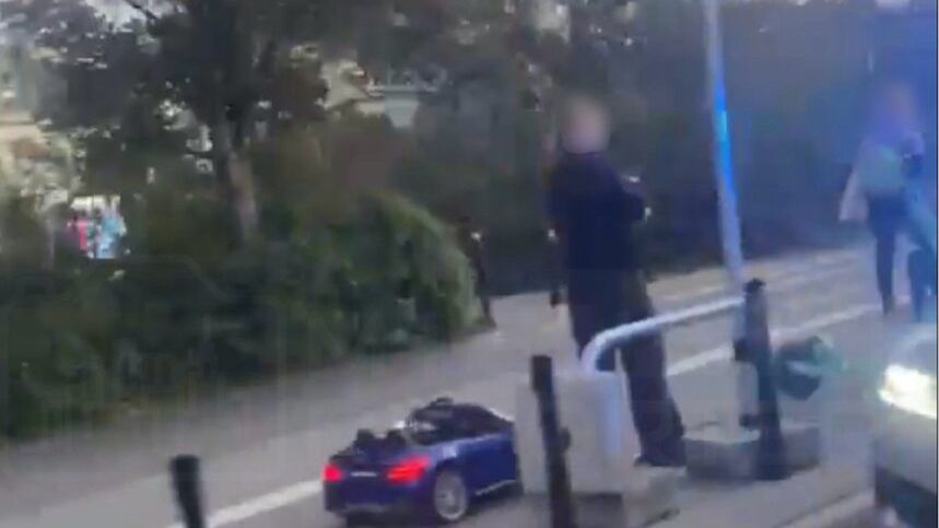 Mężczyzna w czarnym ubraniu obserwujący mały, niebieski samochodzik na chodniku w miejskim otoczeniu.