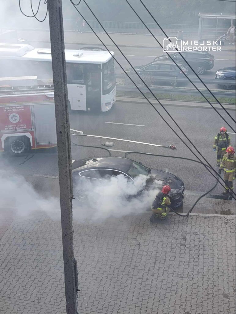 Strażak w hełmie gasi dymiący samochód na ulicy, obok stoi wóz strażacki. W tle widać przejeżdżający autobus.