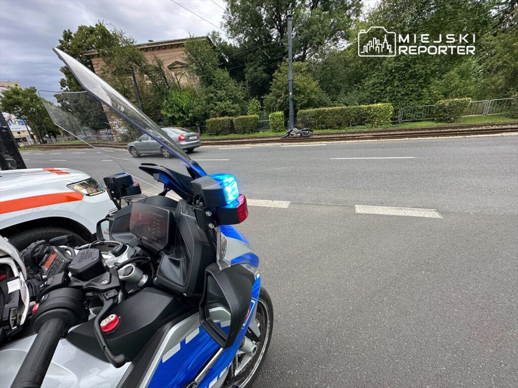 Motocykl policyjny z niebieskim sygnałem świetlnym zaparkowany na ulicy obok radiowozu, w tle zieleń i budynki.