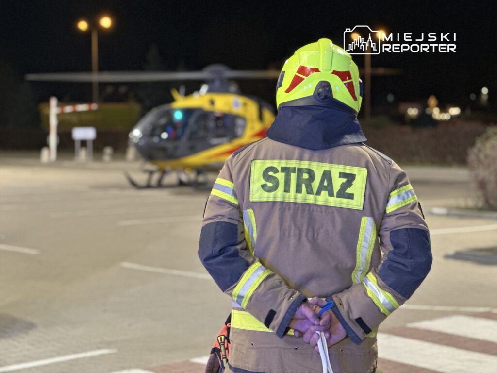 Strażak w mundurze z napisem "STRAŻ" stoi z rękami za plecami przed lądowiskiem śmigłowca ratunkowego nocą.
