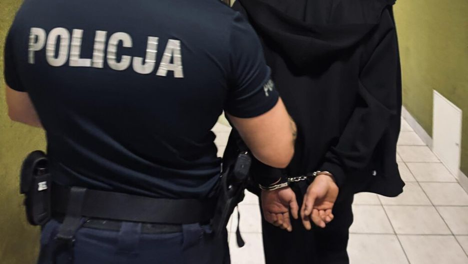 Policjant prowadzi aresztowanego mężczyznę w kajdankach korytarzem w budynku.