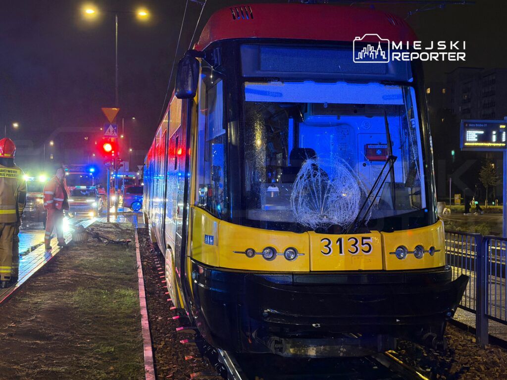 Tramwaj z uszkodzoną szybą stoi na torach, obok grupy ratowników i pojazdów służb ratunkowych w tle.