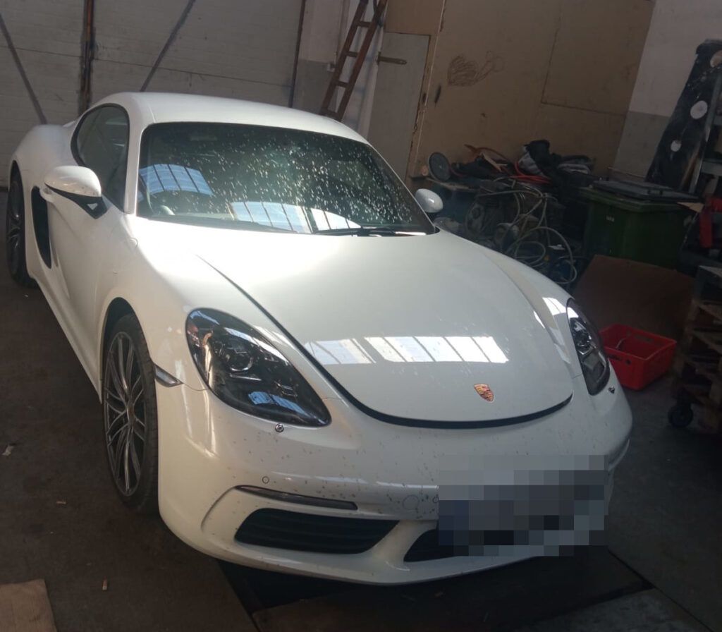 Biały sportowy samochód marki Porsche zaparkowany w warsztacie, otoczony narzędziami i częściami.