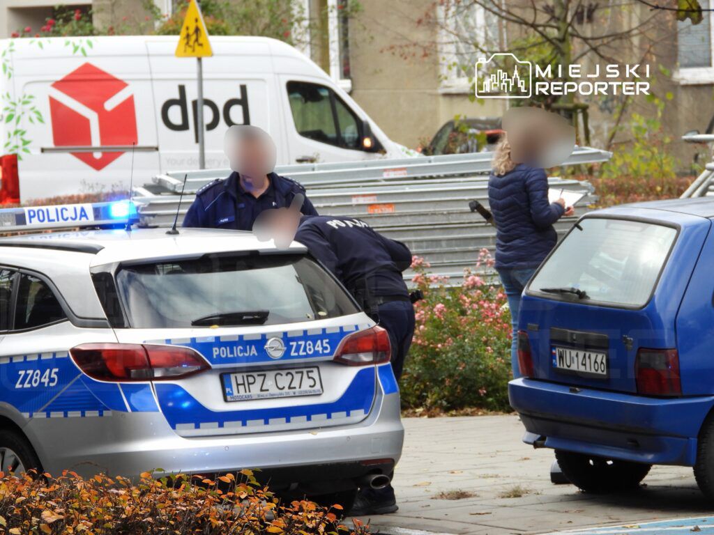 Policjanci w mundurach prowadzą działania przy radiowozie na miejskiej ulicy, obok stoi kobieta z notatnikiem.