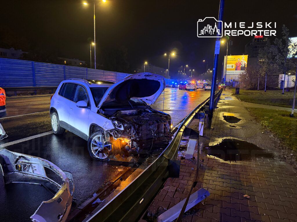 Samochód osobowy z uszkodzoną maską stoi na jezdni, obok leży fragment zderzaka, w tle widać radiowozy policyjne.