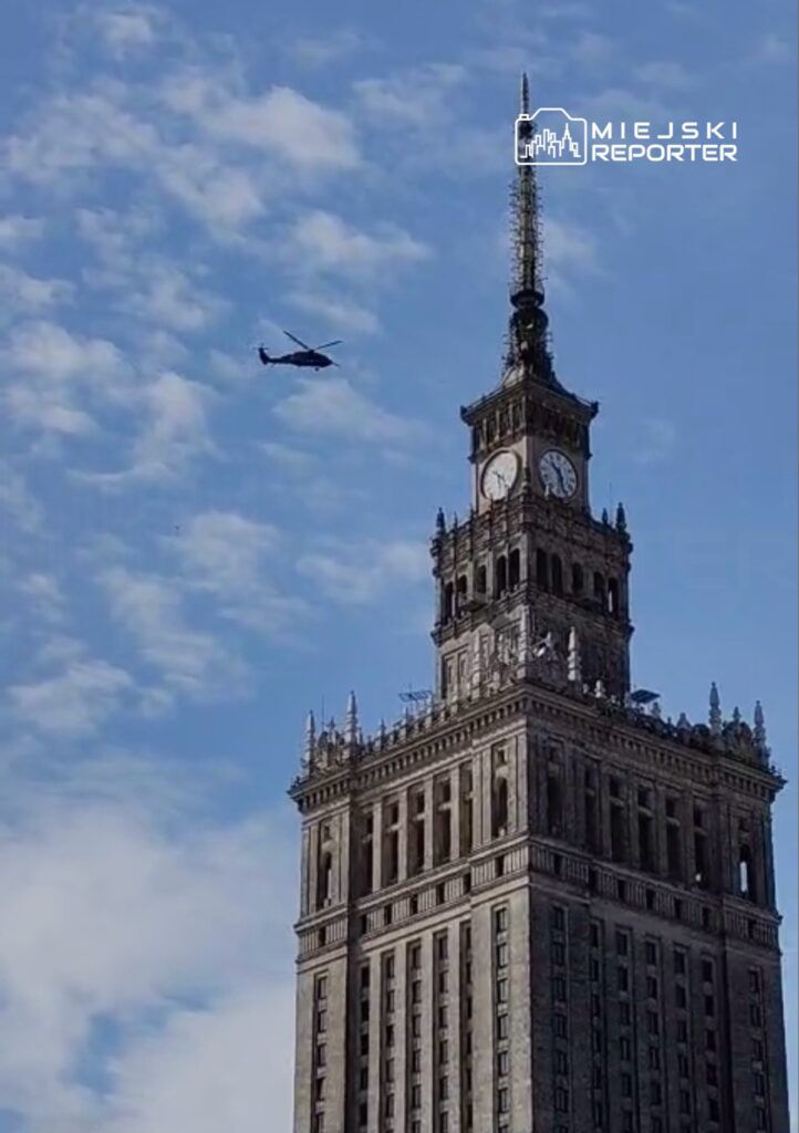 Helikopter przelatujący nad wieżowcem z zegarem w słoneczny dzień, z niebieskim niebem w tle.