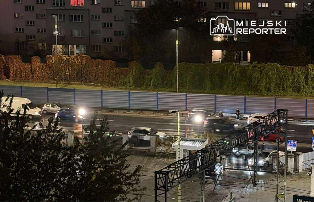 Ruch samochodowy na ulicy w nocy, z widocznymi światłami pojazdów i ogrodzeniem w tle.