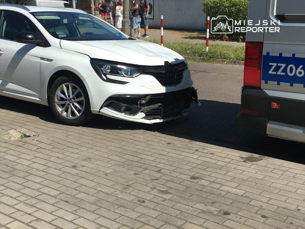 Biały samochód osobowy z uszkodzonym przodem stoi obok pojazdu służb ratunkowych na ulicy.