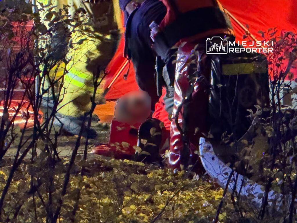 Ratownicy medyczni udzielają pomocy poszkodowanemu w nocy, otoczeni przez krzewy i namioty ratunkowe.