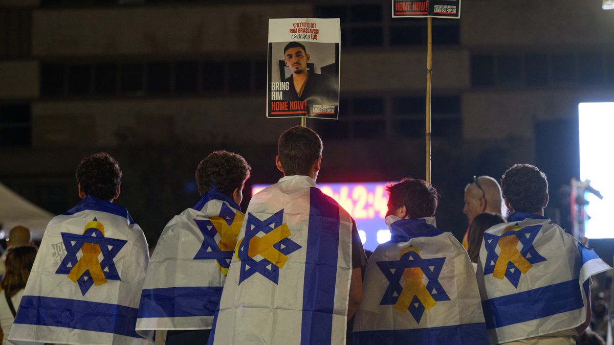TEL AVIV, ISRAEL - OCTOBER 13: Five people draped in Israeli flags with hostage ribbons on them watch tribute videos while waiting for a live stream of the hostage releases to start at Hostages Square early in the morning on October 13, 2025 in Tel Aviv, Israel. The ceasefire deal between Israel and Hamas has brought an end to the two years of war that followed the attacks of Oct. 7, 2023. A condition of the deal was the immediate return of 48 hostages held in Gaza, around 20 of whom were believed to be alive. (Photo by Alexi J. Rosenfeld/Getty Images)