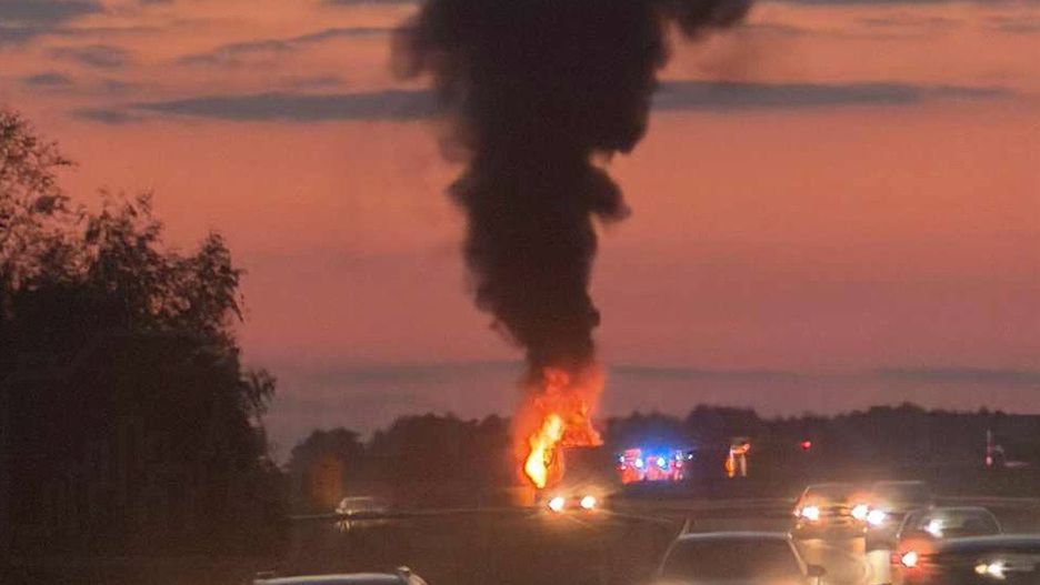 Pojazdy przejeżdżają obok płonącego wraku na drodze, w tle widoczny dym i policyjne światła.