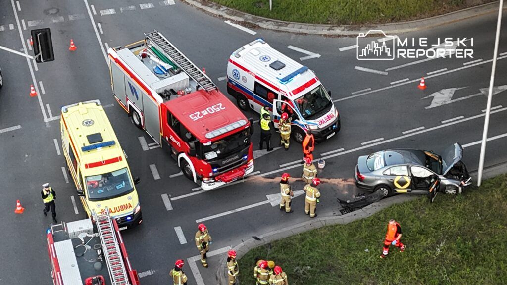 Na skrzyżowaniu stoją samochody ratunkowe; strażacy w hełmach pracują przy rozbitej osobówce.