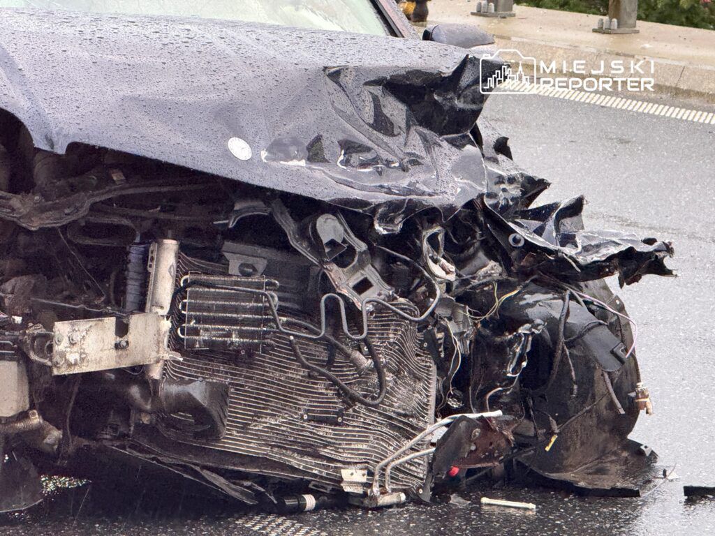 Zniszczony przód samochodu osobowego leżącego na jezdni po wypadku, z widocznymi uszkodzeniami silnika i karoserii.