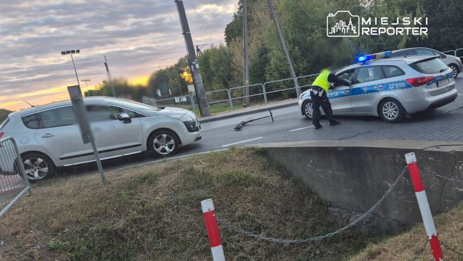 Policjant w kamizelce odblaskowej zabezpiecza miejsce zdarzenia drogowego, obok leży uszkodzona hulajnoga.