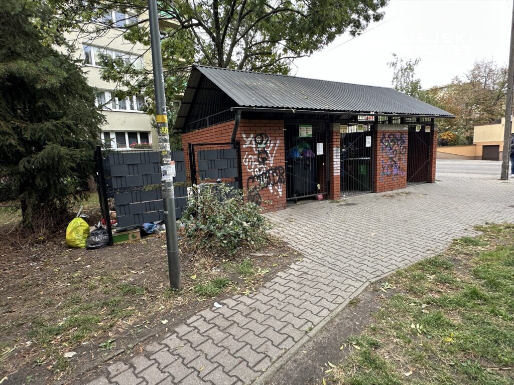 Zabudowana przestrzeń z czerwonej cegły, z zamkniętymi bramami, otoczona graffiti i porozrzucanymi śmieciami.