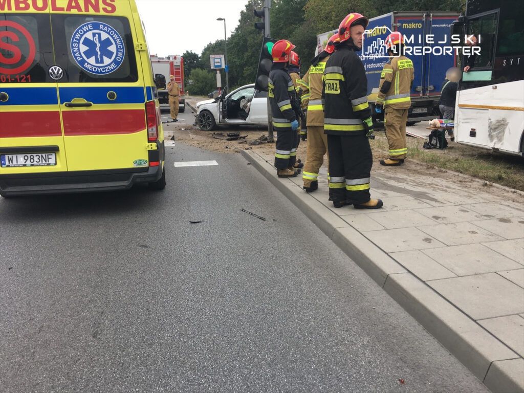 Grupa strażaków w mundurach omawia sytuację obok uszkodzonego samochodu osobowego na drodze. Obok stoi ambulans.
