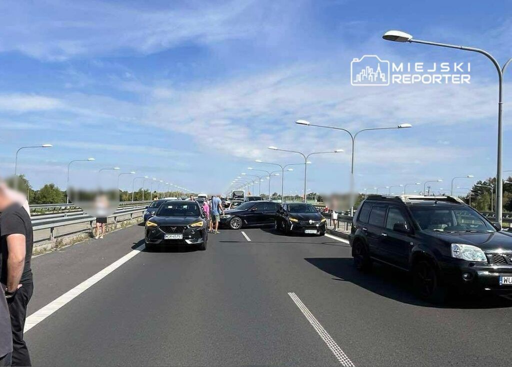 Grupa ludzi stoi na zablokowanej drodze, obok kilku samochodów osobowych, w słoneczny dzień.