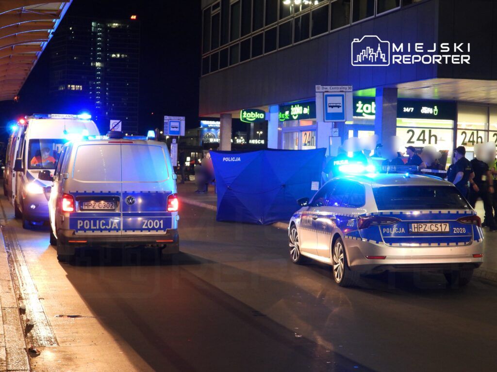 Na nocnej ulicy, policyjny radiowóz i ambulans z włączonymi sygnałami świetlnymi zabezpieczają miejsce zdarzenia.