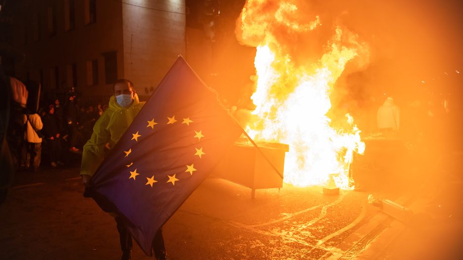 TBILISI, GEORGIA - 2024/11/29: An anti-government protester holds the European flag in front of a makeshift barricade on fire during the demonstration. Following a controversial election last month, ruling party "Georgian Dream" Prime Minister Irakli Kobakhidze announced earlier today that they will no longer pursue a European future until the end of 2028. People gathered on the streets and have been out all night, facing tear gas, water cannons and many arrests. (Photo by Jay Kogler/SOPA Images/LightRocket via Getty Images)