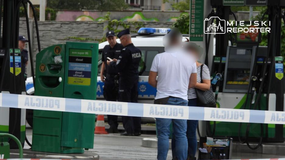 Dwóch policjantów rozmawia z dwiema osobami przy stacji benzynowej, otoczeni taśmą policyjną.