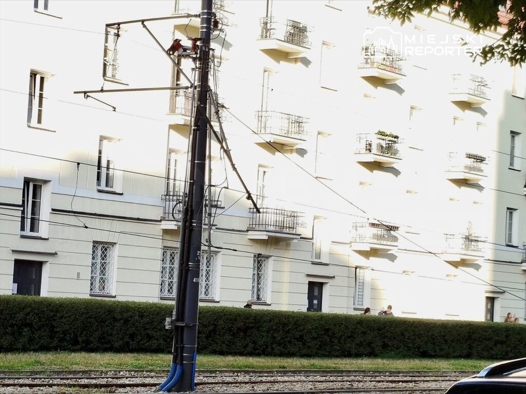 Pracownik na podnośniku wykonuje prace przy linii elektrycznej obok nowoczesnego budynku mieszkalnego.