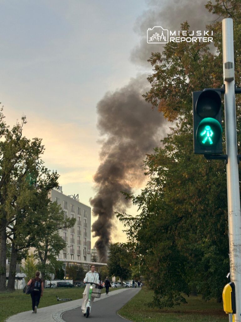 Grupa ludzi spaceruje po ścieżce rowerowej, a w tle unosi się czarny dym z palącego się obiektu. Zielone światło dla pieszych.
