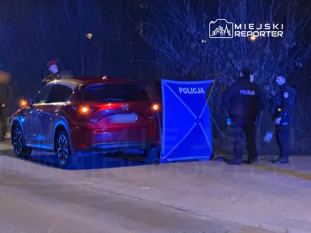 Czerwony samochód osobowy zatrzymany przy drodze, obok policjanci rozmawiający przy niebieskiej barierce z napisem "POLICJA".