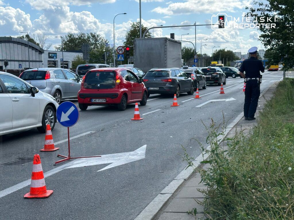Ruch samochodowy wstrzymany na ulicy, policjant kierujący ruchem obok oznakowania i pachołków drogowych.