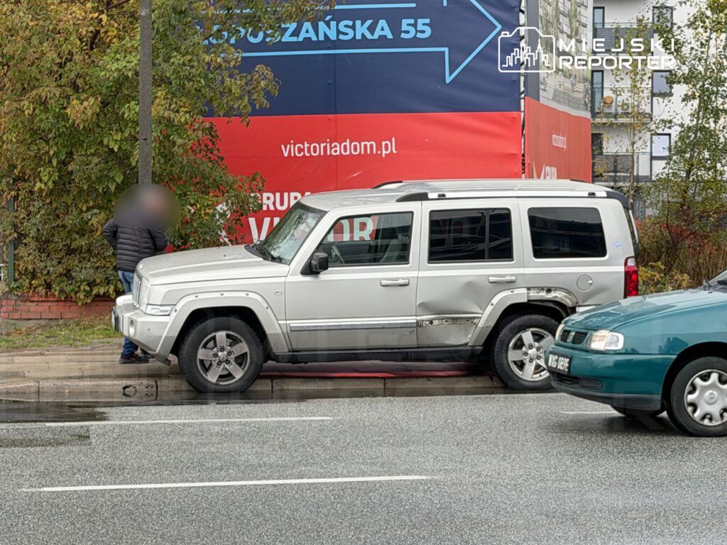 Mężczyzna w czarnej kurtce stoi obok uszkodzonego srebrnego SUV-a na chodniku, w tle widoczny billboard.
