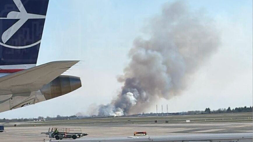 Samolot z logo linii lotniczej na pierwszym planie, w tle widoczny dym unoszący się z pożaru na horyzoncie.