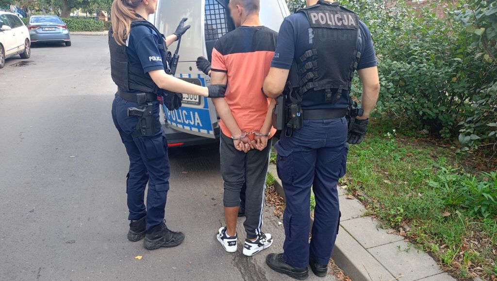 Mężczyzna w kajdankach zatrzymywany przez dwóch policjantów w pobliżu radiowozu na ulicy.
