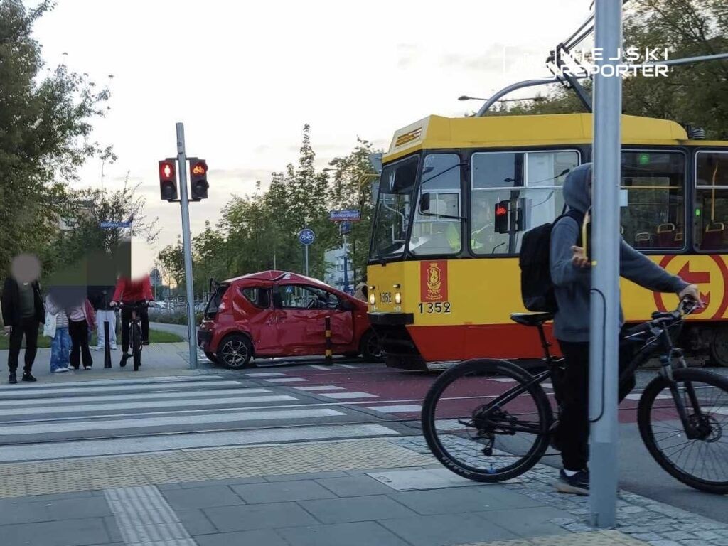 Czerwony samochód osobowy uszkodzony po zderzeniu z tramwajem na skrzyżowaniu, obok przechodnie i rowerzysta.