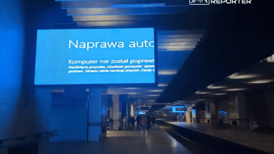 Ekran informacyjny w stacji kolejowej wyświetlający komunikat o błędzie komputera. W tle widać przechodzących pasażerów.