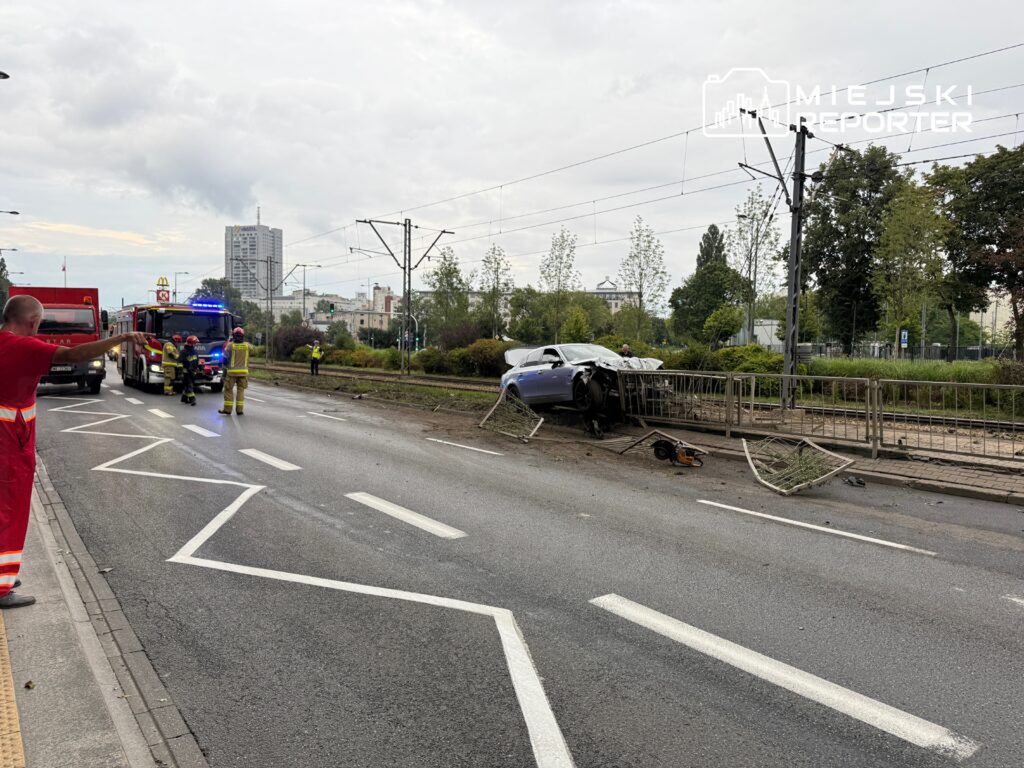 Wypadek samochodowy z uszkodzonym pojazdem na poboczu, obok straży pożarnej i ratowników medycznych w akcji.