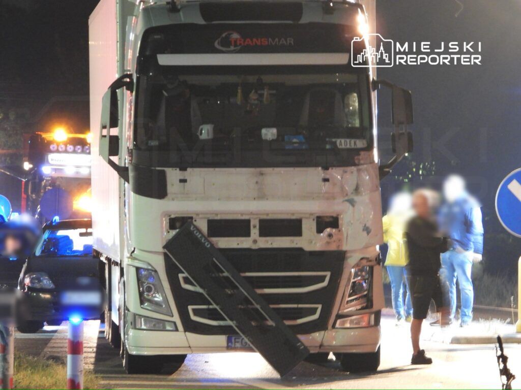 Ciężarówka z naczepą zatrzymana na drodze, obok stoją policjanci oraz kierowca osobowego auta. W tle widoczne światła radiowozu.