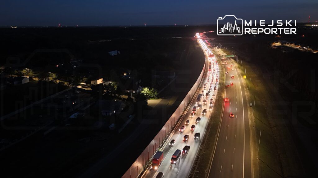 Ruch samochodowy na drodze ekspresowej w nocy, z widocznymi światłami pojazdów i ogrodzeniem po lewej stronie.