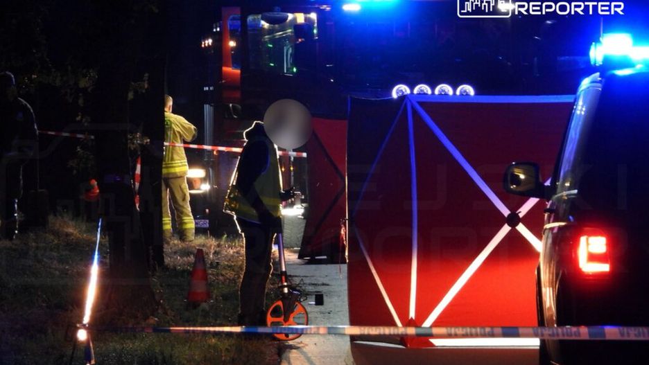 Na zdjęciu przedstawiono akcję ratunkową prowadzoną przez strażaków i policję w nocy. W tle widoczny jest wóz strażacki oraz oznakowane miejsce zdarzenia. Funkcjonariusze zabezpieczają teren i prowadzą działania związane z incydentem drogowym. Widać również sprzęt pomiarowy.