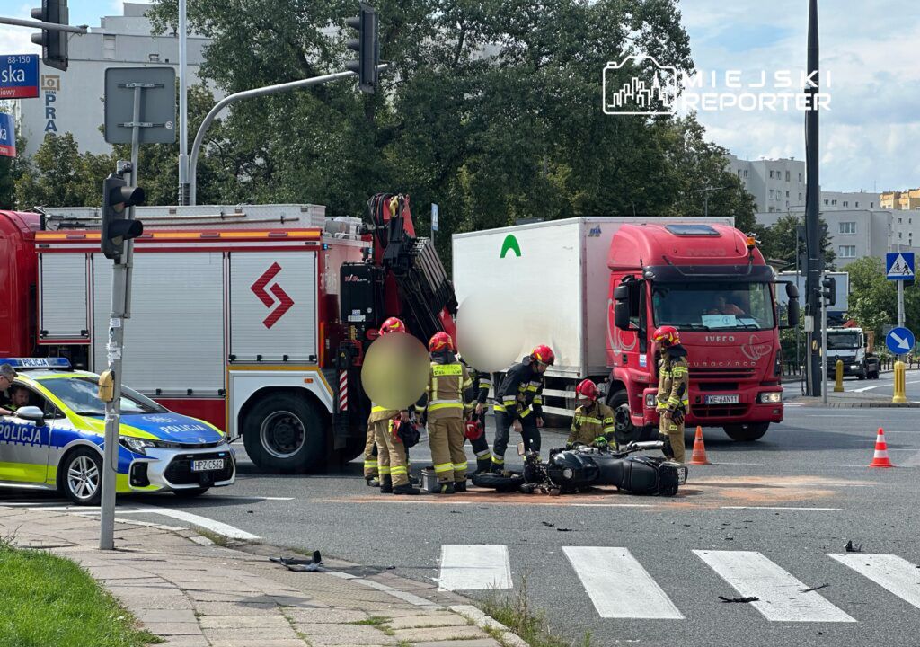 Na skrzyżowaniu ulicy doszło do wypadku drogowego z udziałem motocykla i ciężarówki. Na miejscu interweniują służby ratunkowe, w tym straż pożarna i policja. Widać uszkodzony motocykl oraz zaparkowane pojazdy służbowe. Ruch w okolicy jest utrudniony.