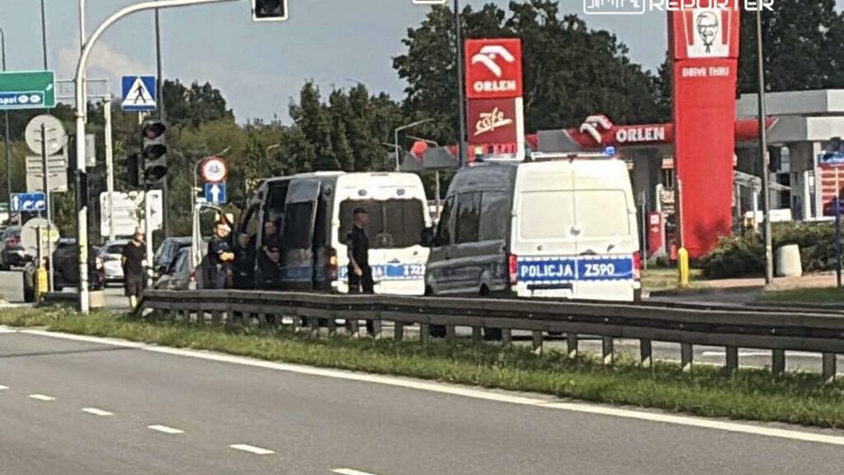 Grupa policjantów w mundurach prowadzi działania przy zatrzymanych pojazdach obok stacji benzynowej.