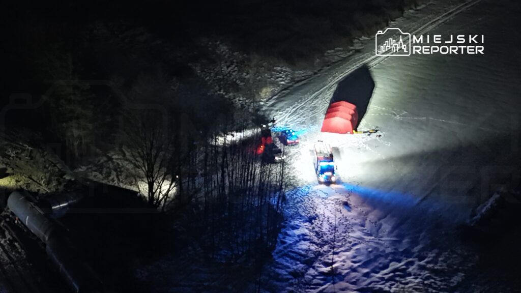 Służby ratunkowe pracują w nocy w terenie pokrytym śniegiem, obok czerwonego namiotu i pojazdów z włączonymi sygnalizatorami.